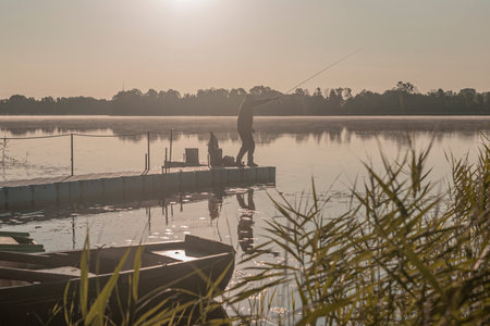 Fisherman fishing in foggy morning. Man silhouette resting with rod on river or lake in mist.