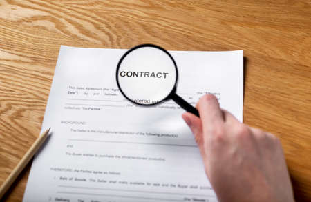 Paper contract on wood desk with magnifying glass in female hand.