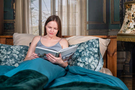 Woman lying in bedon pillows and reading book, resting at home in the evening.