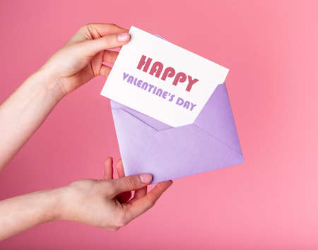 Hands putting card with happy Valentines day greeting in envelope. Celebration postcard on pink background. High quality photoの写真素材