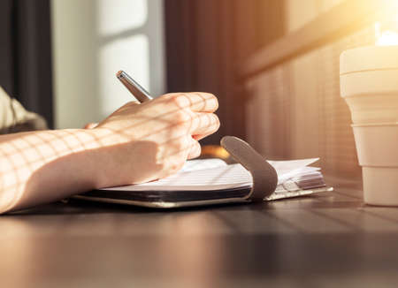 Female hands closeup taking notes in notebook. Woman sitting at table with collapsible silicon mug...