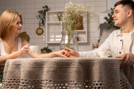 Happy couple sitting at kitchen table, drinking tea or coffee, holding hands, looking at each other....