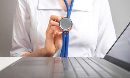 Doctor holding stethoscope while sitting at desk with laptop. Woman in lab coat treating patients. Healthcare and medicine concept. High quality photoの写真素材