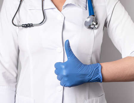 Doctor with thumb up gesture. Good health, trust in medical professional competence concept. Woman in lab coat and in gloves showing approval sign with hand. High quality photoの写真素材