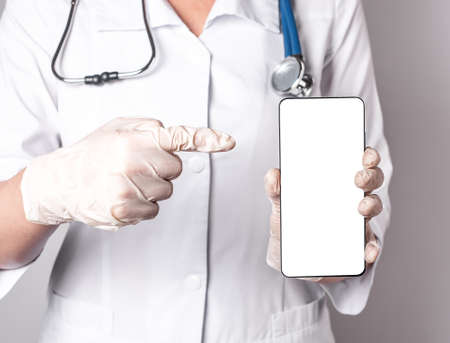 Doctor forefinger pointing to phone screen mockup. Online patient treatment, using medical application in smartphone, telehealth concept. Woman in lab coat with stethoscope and gloves. High quality photoの写真素材