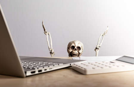 job burnout. Skeleton peeking out from desk with laptop and calculator. computer geek. Emotional and physical exhaustion, frustration, stress from work overload, deadlines. high quality photoの写真素材