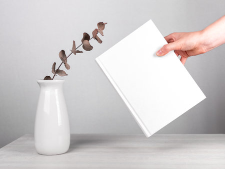 Hand holding book mockup near flowers in vase at table. Minimalist style. Reading leisure, harmony concept. Novel with white cover. High quality photoの写真素材
