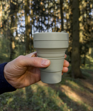 Hand holding silicon collapsible cup, coffee mug in sunny forest, nature. Eco and zero waste...