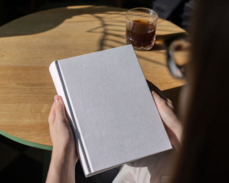 Book cover mock up, hand holding blank clean hardback mockup on wooden deskの写真素材