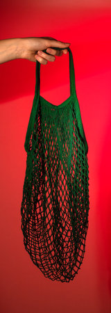 Mesh bag in hand, reusable cotton fabric for eco-friendly shopping. Sustainable grocery sack, biodegradable string package, organic lifestyle concept. Red background.の写真素材