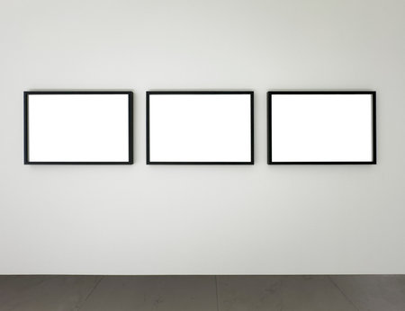 Three black rectangular picture frames mounted on a white wall above a gray floor, modern gallery mockup for displaying photography, posters, or digital artwork in a clean minimalist interior.の写真素材