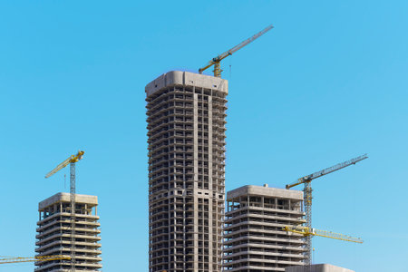 Construction of tall multistorey residential tower in developing metropolitan district. Unfinished concrete and steel framework with cranes under clear blue sky. City architecture, housing, propertyの写真素材