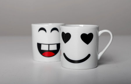 Two white ceramic mugs with playful emoji faces. Cute smiling happy and valentine love cup designs,...