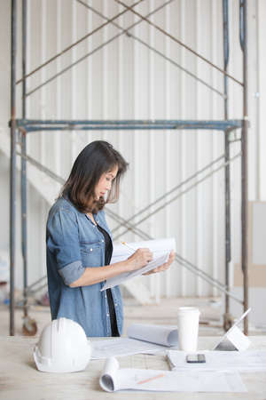 Beautiful Asian engineer woman in blue jeans shirt standing and writing, white safty helmet on desk.の写真素材