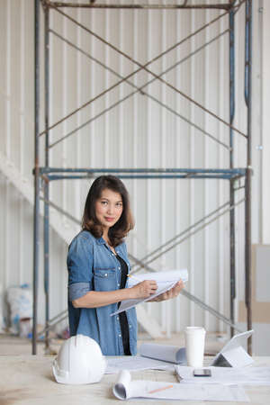 Beautiful Asian engineer woman in blue jeans shirt standing, writing and looking, white safty helmet on desk.の写真素材