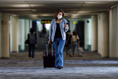 Beautiful Asian traveler wearing hygiene protective mask holding luggage bag walking and using...