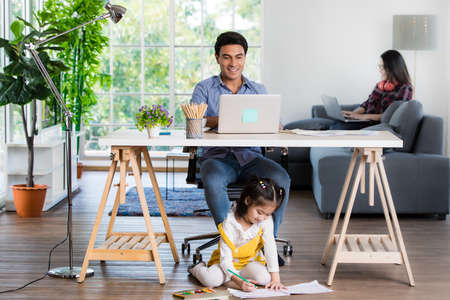 Mixed race family sharing time in living room. Caucasian father using notebook computer to work and half-Thai playing and painting under desk while Asian mother with laptop working her job on sofa.の写真素材