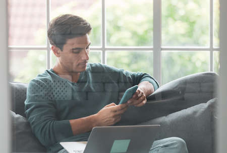 Young and handsome Caucasian businessman wearing casual cloth sitting happily on sofa and using ...