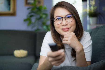 Cute Asian woman laying on sofa with relax and cozy gesture holding remote control aiming to tv,...