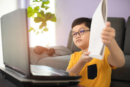 Young and cute 10s little Asian schoolboy wearing eyeglassed showing his homework for teacher during online studying by using laptop notebook computer in living room at home. Distance education idea.の写真素材