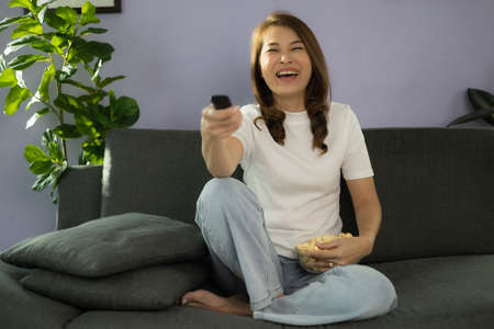 Cute Asian woman sitting on sofa with relax and cozy gesture holding remote control, watching...