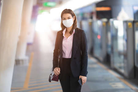 Asian female passenger in a black suit wearing medical hygiene protective mask standing alone waiting for train at station. Concept for businesspeople prevents health from coronavirus or COVID-19.の写真素材