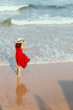 Woman photographer in red dress  standing  and soaking in water on beach and taking photo to wide and far point of the sea. Blues sky background and copy space. Idea for lifestyle of modern woman.の写真素材