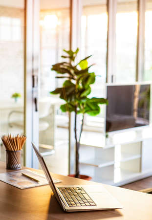 Notebook computer on desk in living room with ray of warm orange sunlight shine from windows and nobody in house. Idea for work at home or home base working of modern business lifestyle.の写真素材