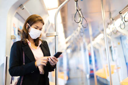 Bueatiful adult Asian businesswoman passenger commuter wearing medical hygiene protect mask. Standing and using cell phone inside empty subway or sky train car. Idea for abandoned city from COVID-19.の写真素材