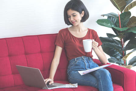 Young and beautiful short hair half Thai - German girl holding coffee cup sitting on red sofa and using a laptop computer to work from home with a smiling face and happiness.の写真素材