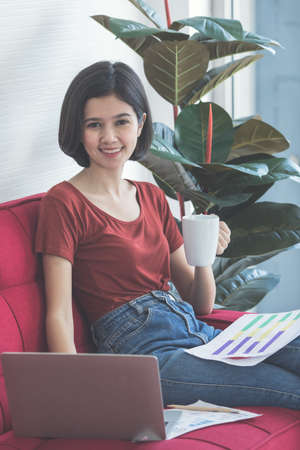 Young and beautiful short hair half Thai - German girl holding coffee cup sitting on red sofa and using a laptop computer to work from home with a smiling face and happiness.の写真素材