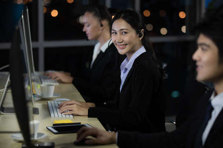 Beautiful Asian young pretty woman wearing headphone headset working overtime late at night in call center office with her colleagues' team and looking to camera with smile face. Idea for hard job.の写真素材