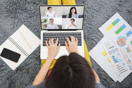 Businesswoman in casual dress sitting on floor beside sofa in living room with work paper, using laptop notebook computer to meeting with four other friends. They are happy and laugh with funny.の写真素材