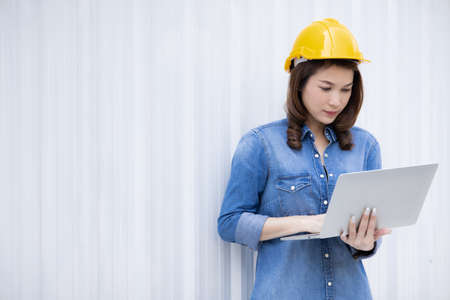 Beautiful Asian female engineer in jeans dress wearing yellow safety hard hat using laptop computer notebook doing job at construction site outside office. Idea for modern working woman.の写真素材