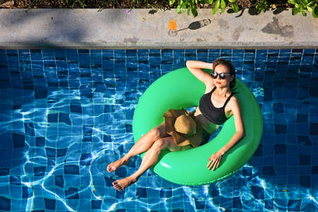 Top view shot woman with big hat laying on balloon in blue water swimming pool with copy space,...