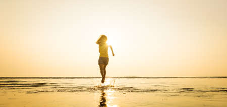 Woman running from beach into seawater with happiness and excited feeling when seeing morning sunlight over skyline and beautiful blue sky. Using high speed shutter to stop splash of water drops.の写真素材
