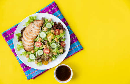 Top view shot of chicken salad  with several kinds of fresh vegetable in white plate on yellow background and cloth. Copy space on right.の写真素材