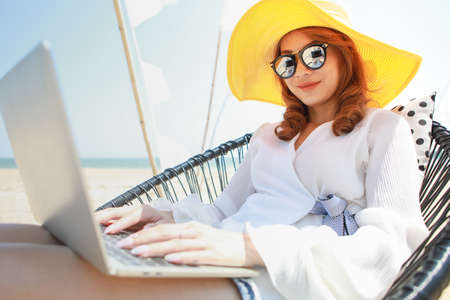 Beautiful woman wearing yellow hat sitting on chair and using laptop computer near beach in sunny day with relaxing pose. Idea for lifestyle of modern woman who can work and life balanced.の写真素材