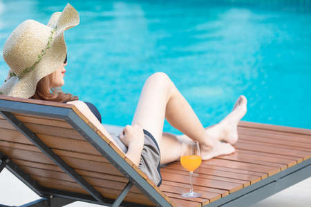 Woman wear big hat lay on wood bed beside swimming pool with blue water and looking to somewhere. There is glass of orange juice near her.の写真素材