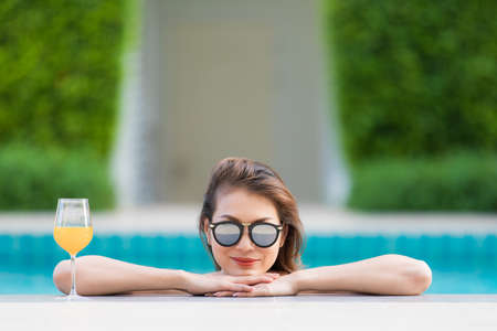 Beautiful Asian woman wear bikini and sunglasses standing in blue water pool with relax manner, looking to camera with glass of orange juice.の写真素材