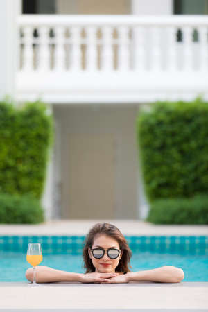 Beautiful Asian woman wear bikini and sunglasses standing in blue water pool with relax manner, looking to camera with glass of orange juice.の写真素材
