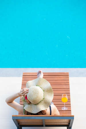 Woman wear big hat lay on wood bed beside swimming pool with blue water. There is glass of orange juice near her.の写真素材