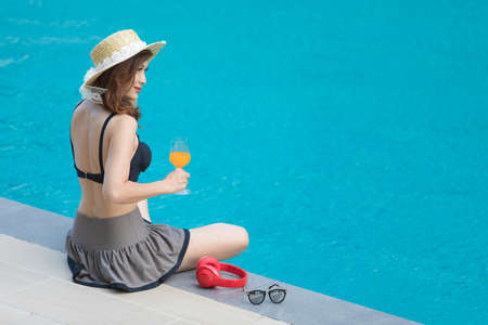 Woman wear big hat sitting  beside blue water swimming pool with red headphone and sunglasses, looking to somewhere. There is glass of orange juice in hand.の写真素材