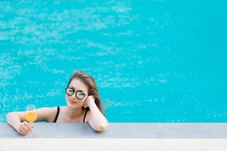 Beautiful Asian woman wear bikini and sunglasses standing in blue water pool with relax manner, looking to camera with glass of orange juice.の写真素材