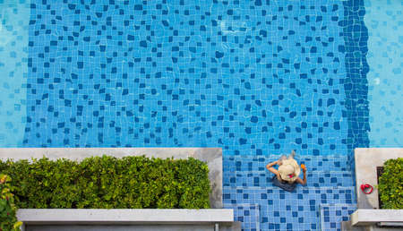 Woman wear big hat  sitting in swimming pool holding glass of orange juice, taken from top view angle.の写真素材