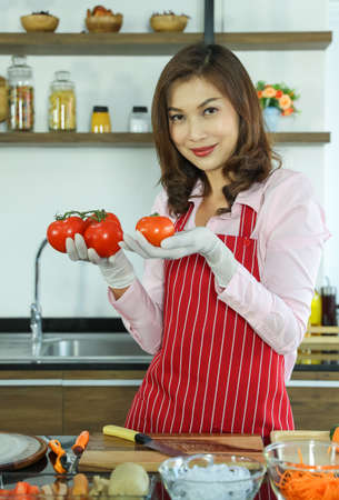 Matue adult Asian female housewife in apron standing in kitchen holding fruit ready to make a vegetable healthy salad and looking to camera.の写真素材