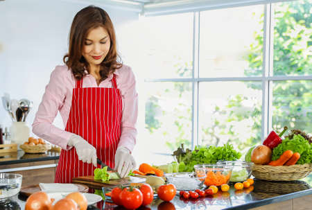 Beautiful mature adult Asian female housewife in apron standing in kitchen with various kinds and colorful vegetables and fruit preparing and making salad rolls. Concept of vegan and healthy food.の写真素材
