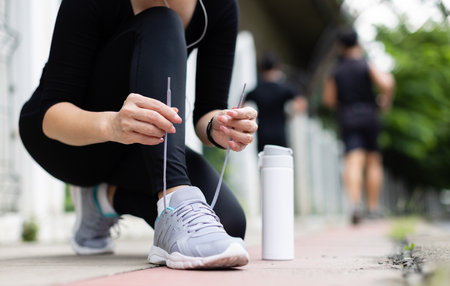 Crop unrecognizable woman runner wearing hygiene protective face mask sitting and tieing shoe rope during workout running in city park. Concept of healthcare in time of coronavirus outbreak.の写真素材