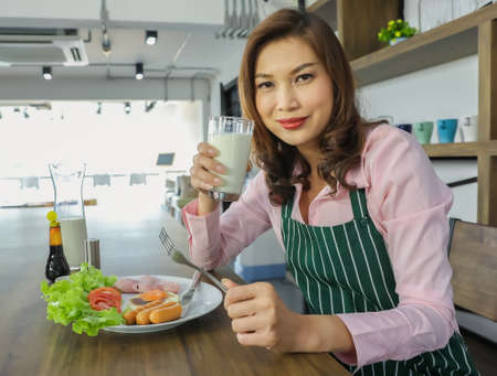 Matue adult Asian female housewife in apron sitting at table in kitchen holding glass of milk and ready to eat breakfast. Happy cute woman starting new day in morning with delicious food.の写真素材