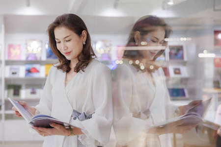 Portrait of Asain pretty cheerful female standing and reading book in library and lean on glass wall with happy, focus and concentrate with reflection in clear mirror.の写真素材